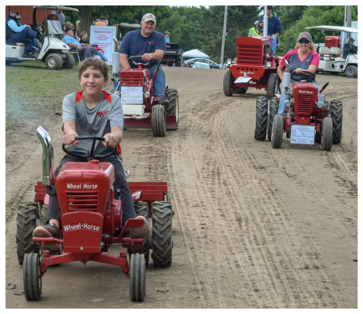 A group of people riding red tractors down a dirt road near their home.